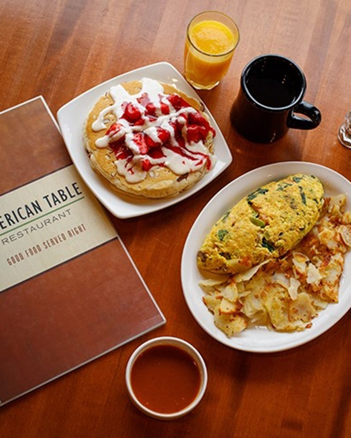 An image of an omelet, hashbrowns, stack of pancakes and beverages on a dining table next to a menu at American Table Diner in Princeton, IN